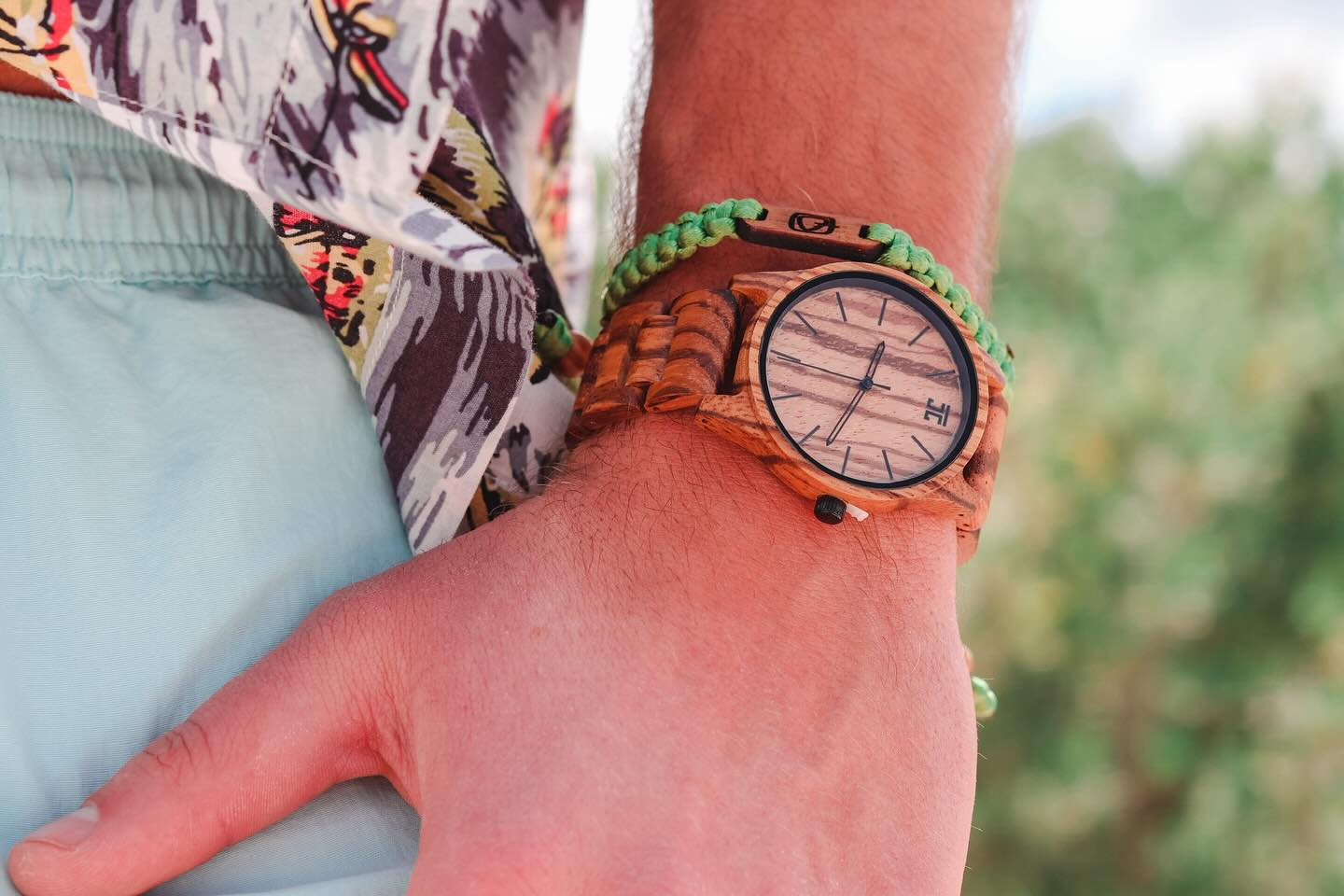 Close-up of a wrist wearing a handmade wooden watch with striped wood grain and green braided bracelet outdoors