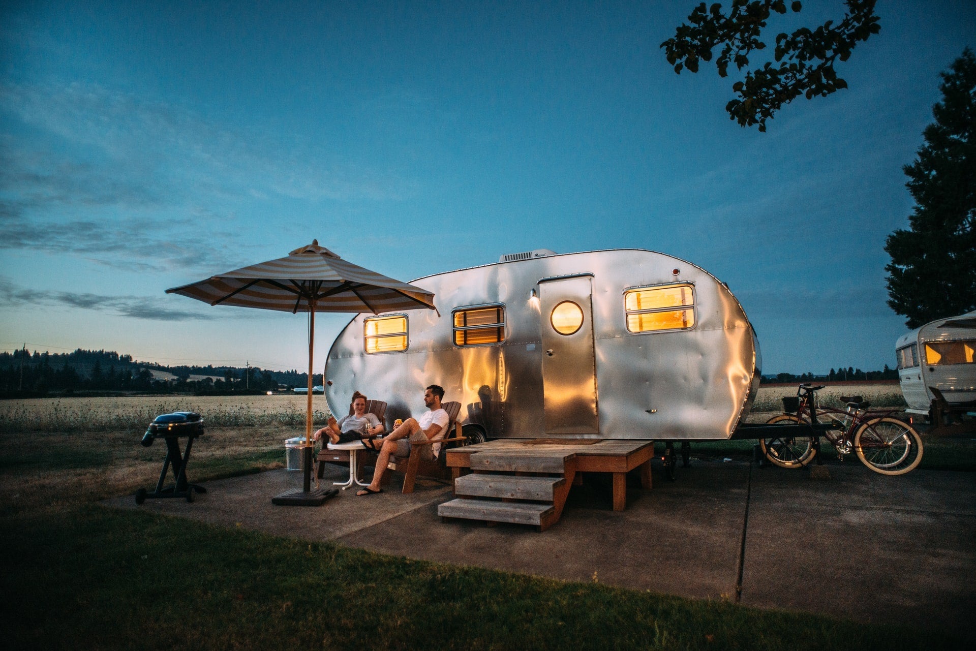 Couple relaxing outside shiny silver camper trailer at dusk with umbrella and barbecue grill
