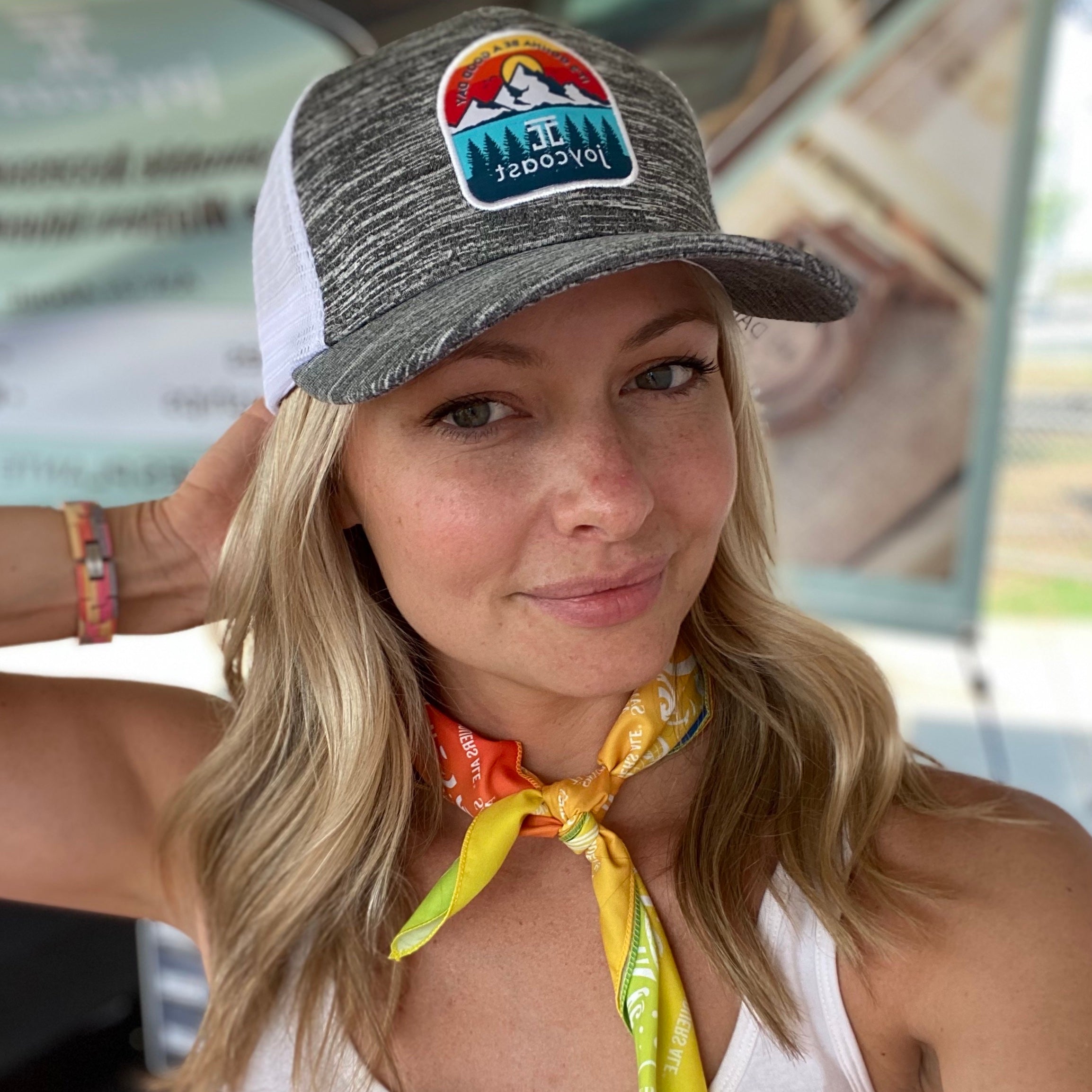 Blonde woman wearing grey and white Joycoast cap with mountain logo and a colorful neck scarf
