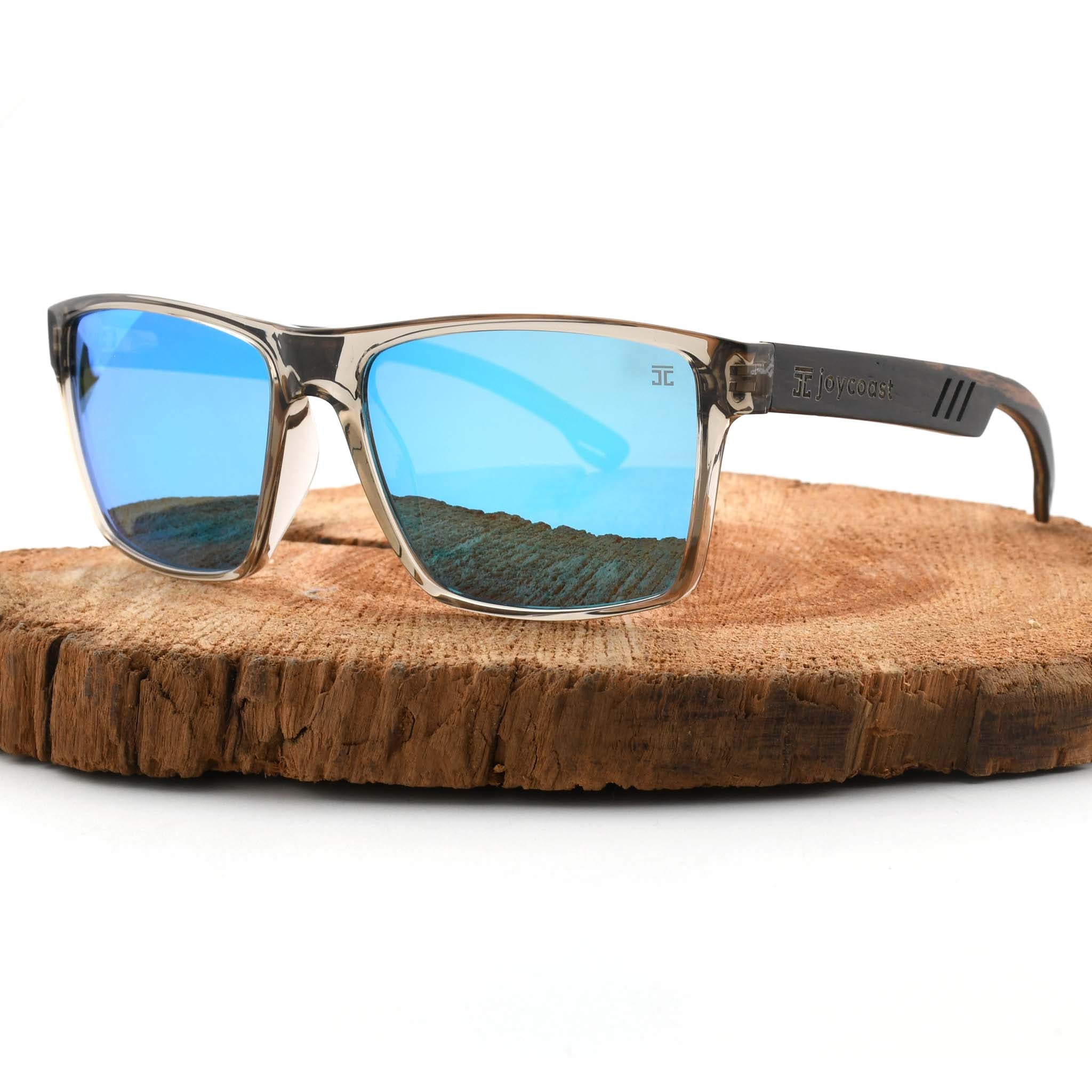 Joycoast Apex transparent frame sunglasses with blue reflective lenses and wooden arms, displayed on a natural wood slice.