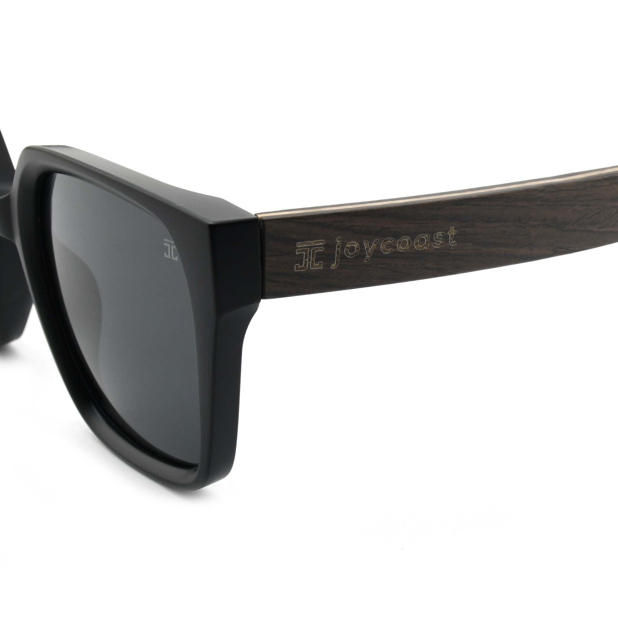 Close-up of a black wooden sunglasses arm with the "Joycoast" logo engraved near the hinge.