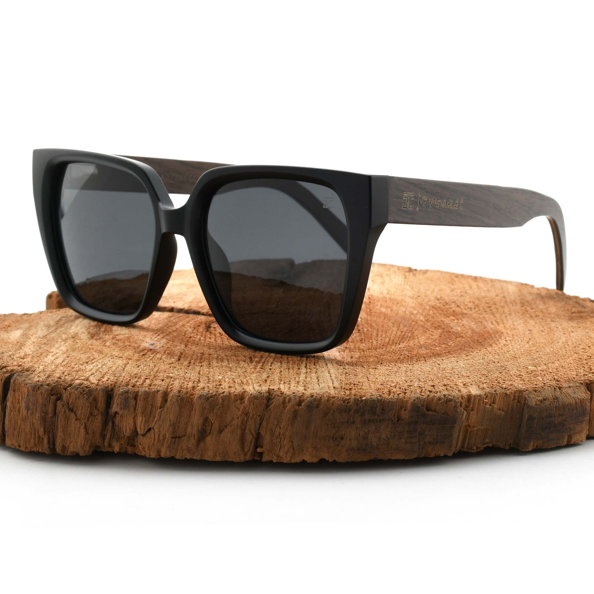 Joycoast black wooden sunglasses with dark lenses, displayed on a natural wood slice.