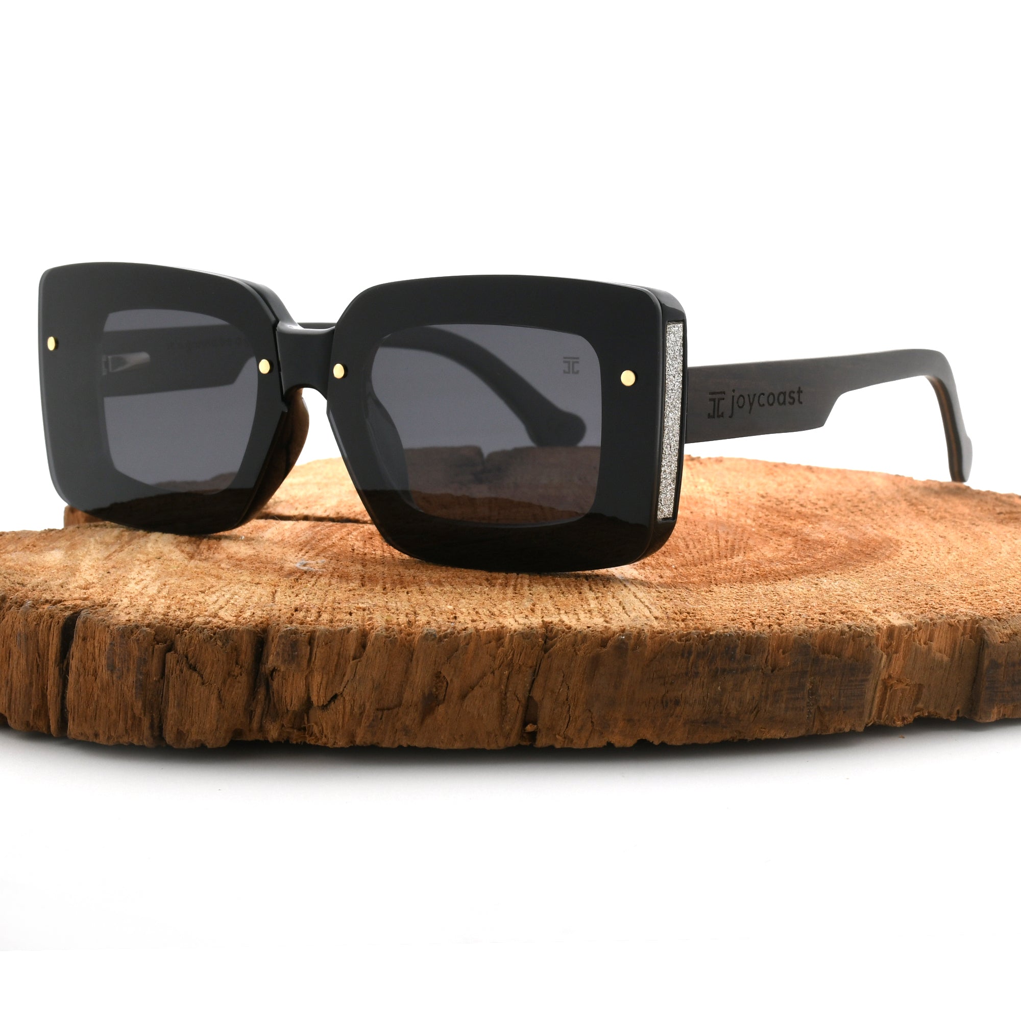 Joycoast black wooden square sunglasses with dark lenses, displayed on a natural wood slice.