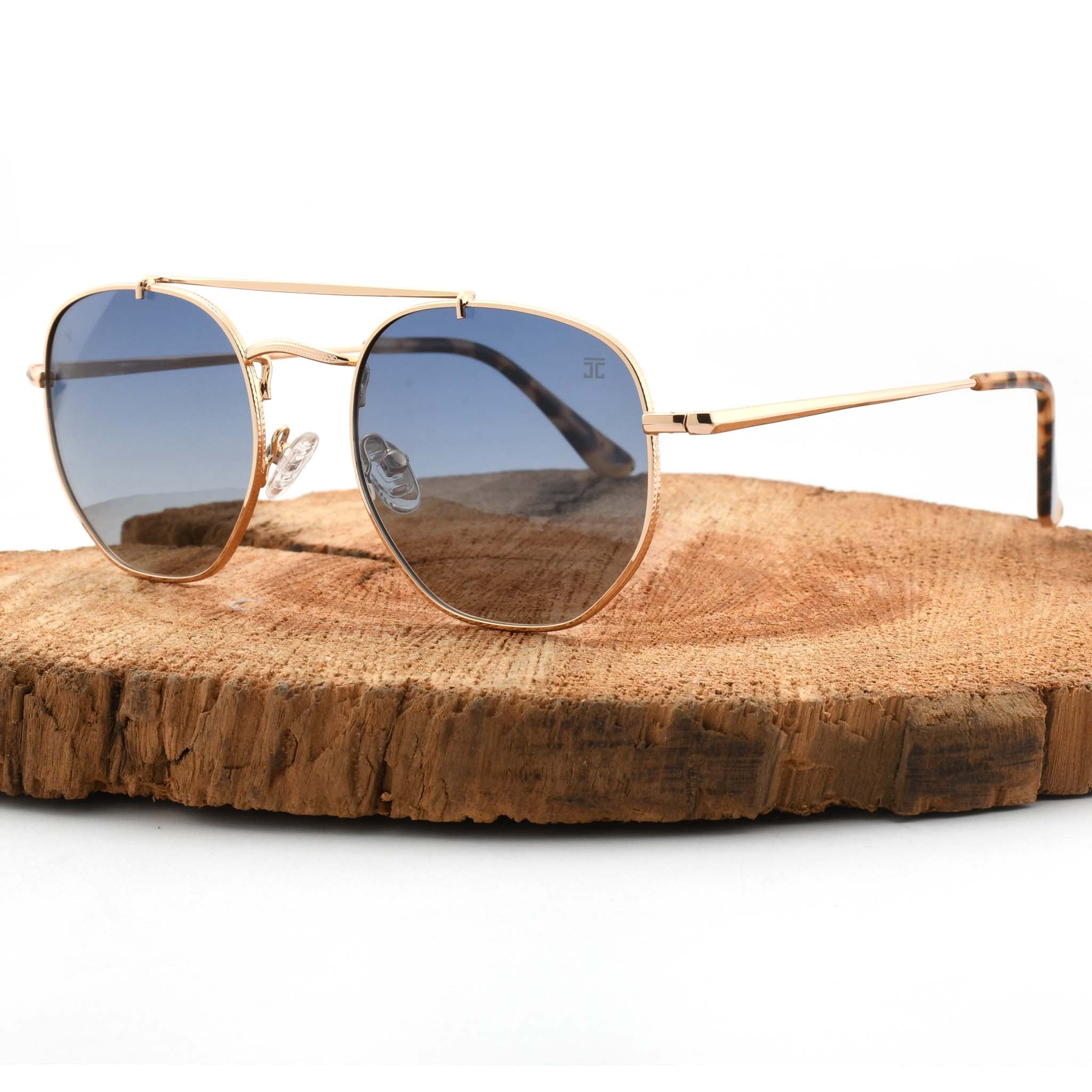 Joycoast Skyline gold metal sunglasses with light blue gradient lenses, displayed on a natural wood slice.