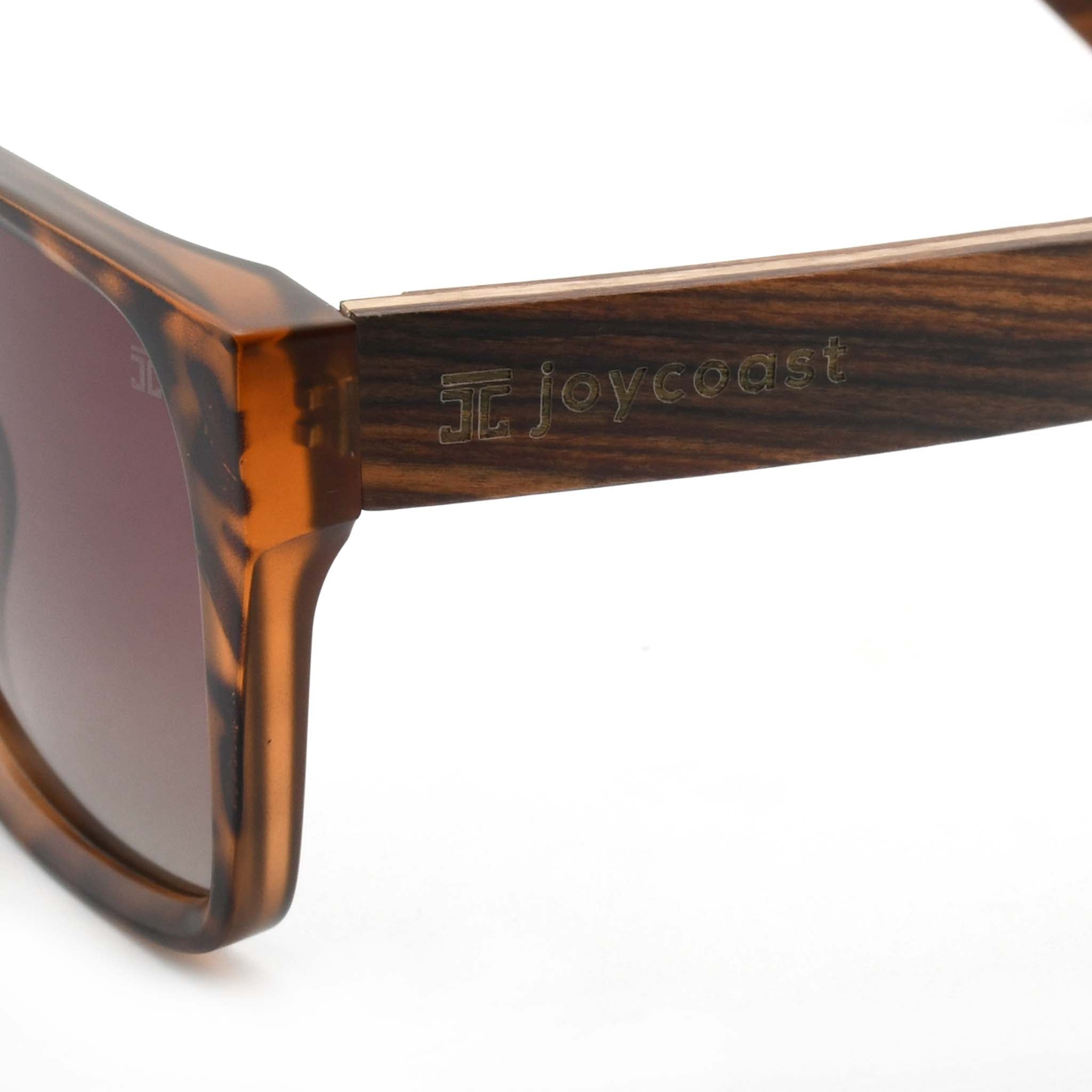 Close-up of a wooden sunglasses arm with the "Joycoast" logo engraved near the hinge of the tortoise shell frame.
