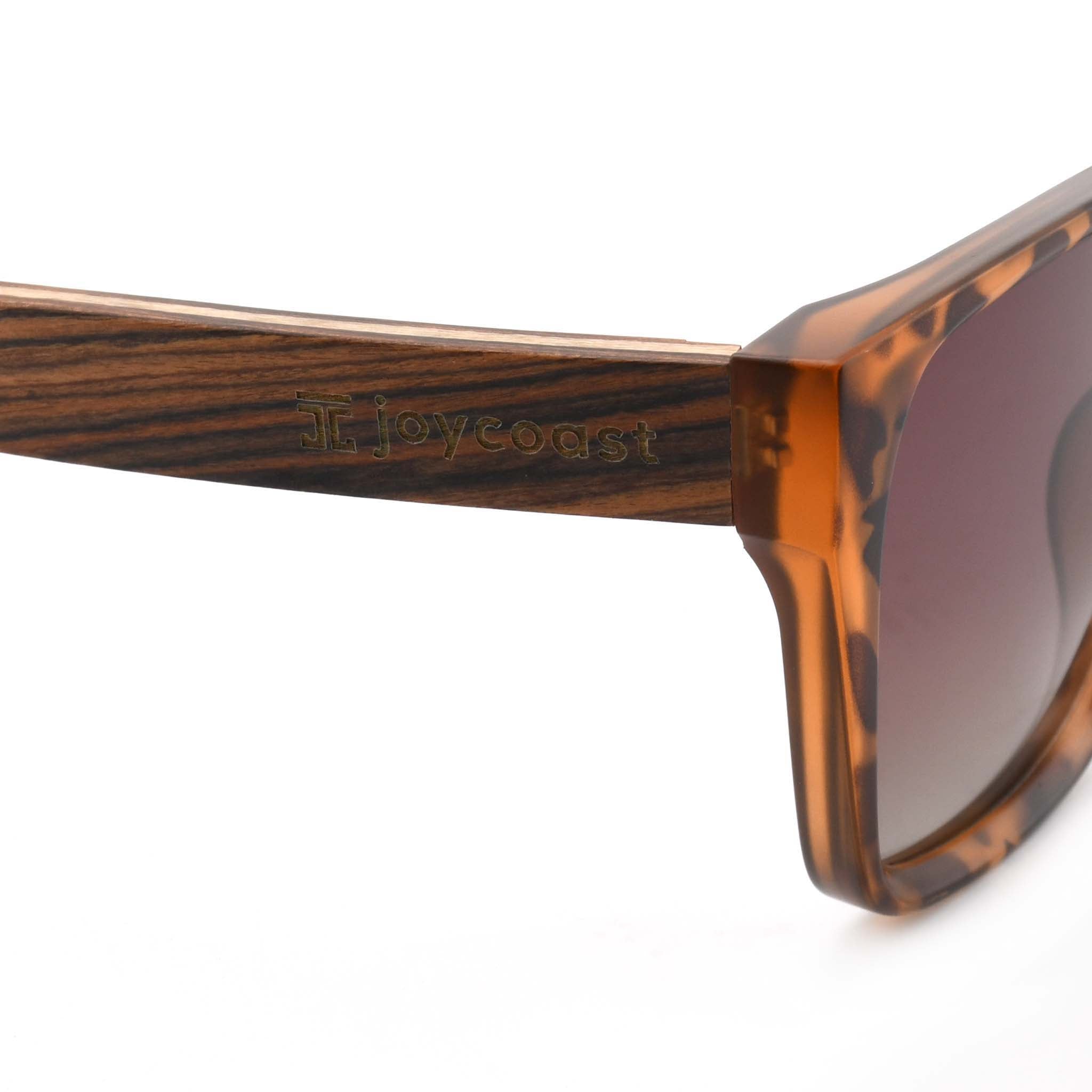 Close-up of a wooden sunglasses arm with the "Joycoast" logo engraved near the hinge of the tortoise shell frame.