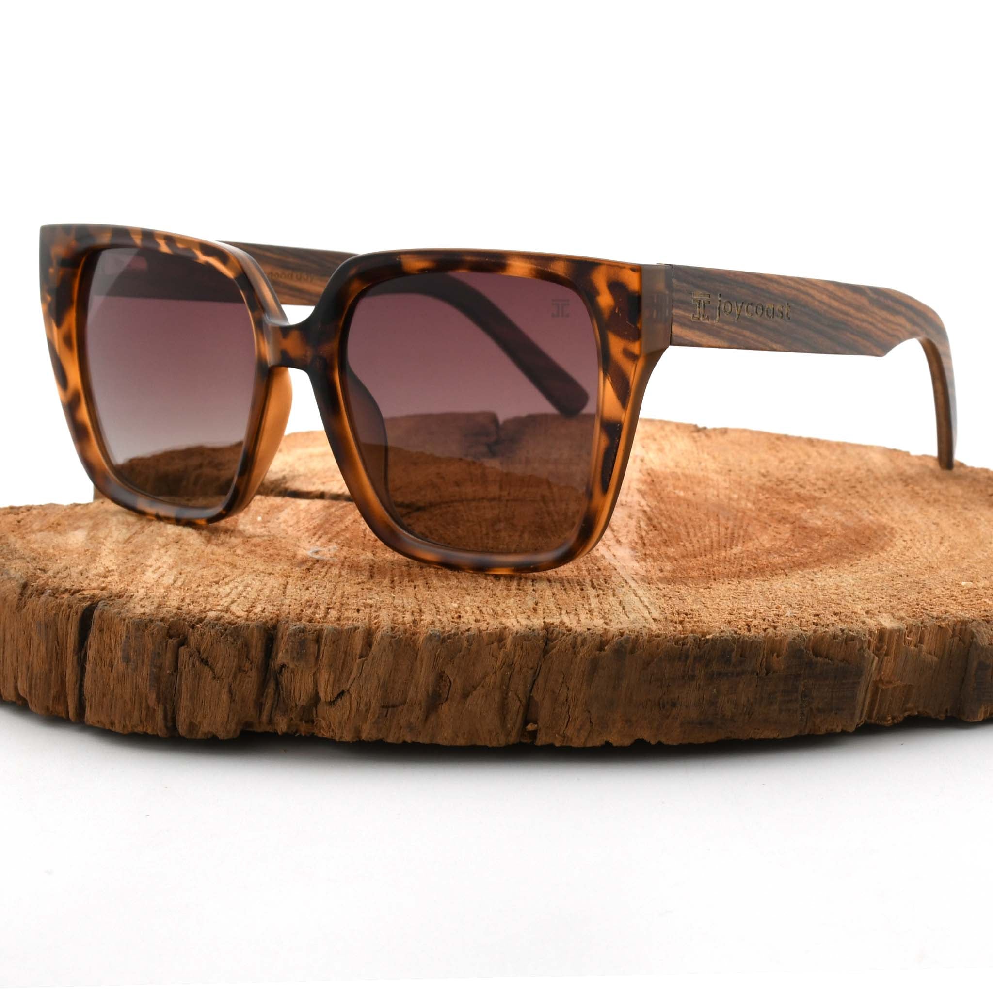 Joycoast tortoise shell sunglasses with brown gradient lenses and wooden arms, displayed on a natural wood slice.