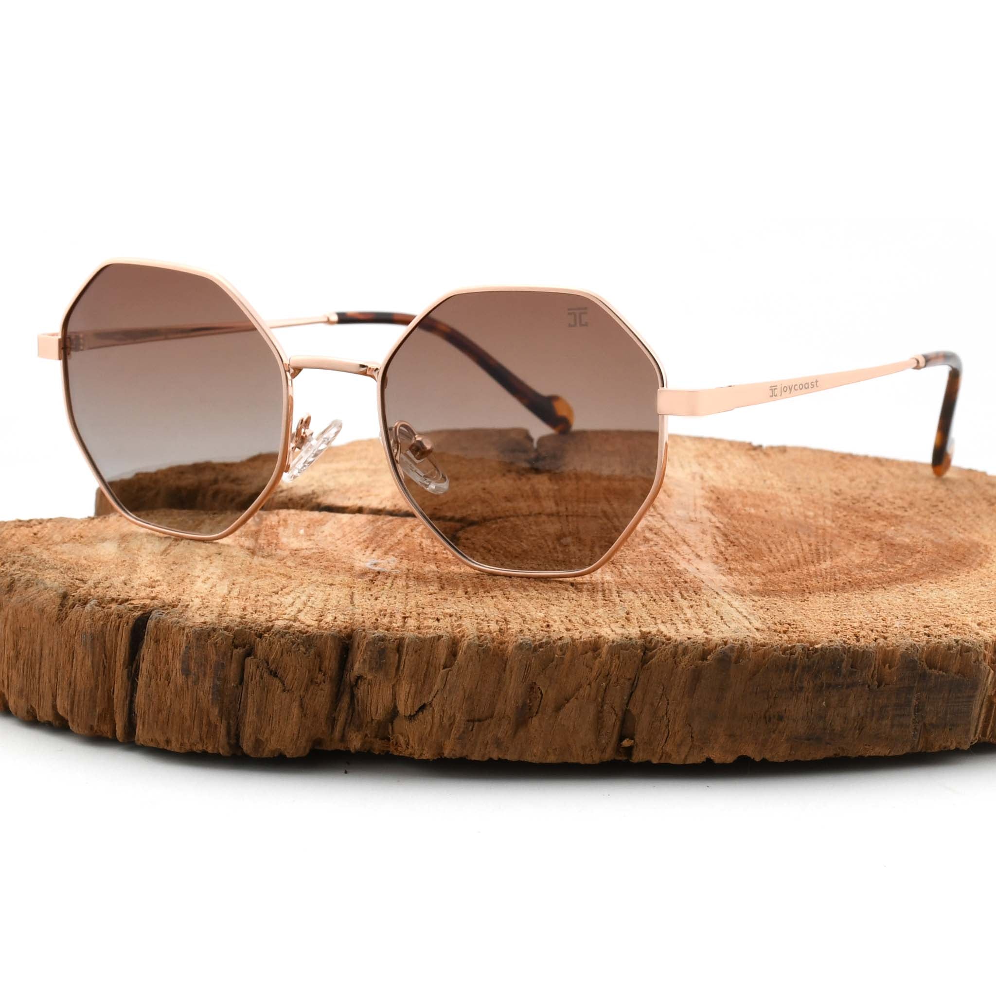 Joycoast Vesuvio roose gold metal hexagon-frame sunglasses with brown/red gradient lenses, displayed on a natural wood slice.