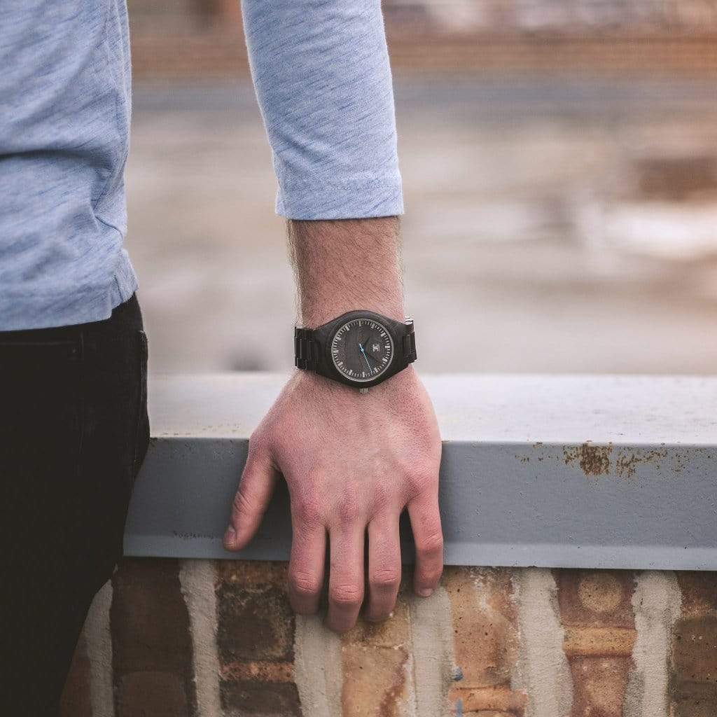Close-up of a person's wrist wearing a black wooden watch by Joycoast, resting on a stone ledge outdoors
