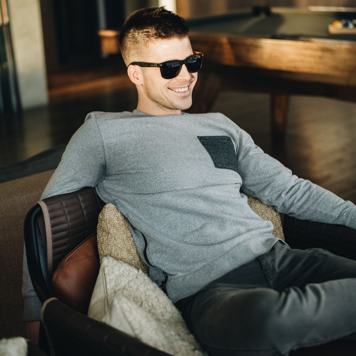 A man wearing sunglasses and a sweatshirt sits casually on a chair, exuding a relaxed demeanor.