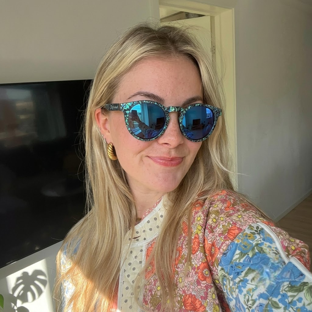 A woman wearing Joycoast sunglasses and a floral shirt, smiling outdoors in bright sunlight.