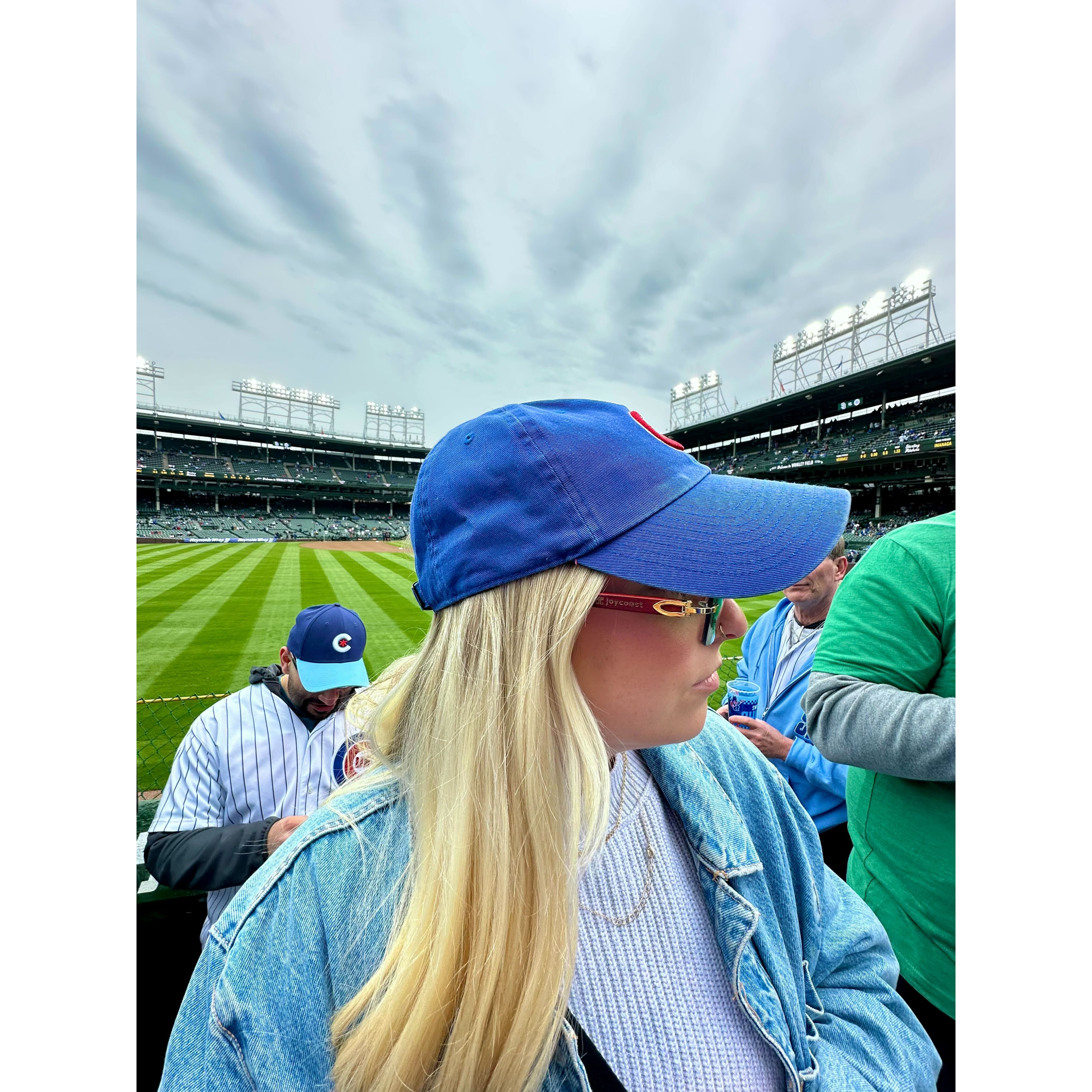 A blonde woman wearing a blue cap and denim jacket, looking to her right, with a baseball stadium in the background.