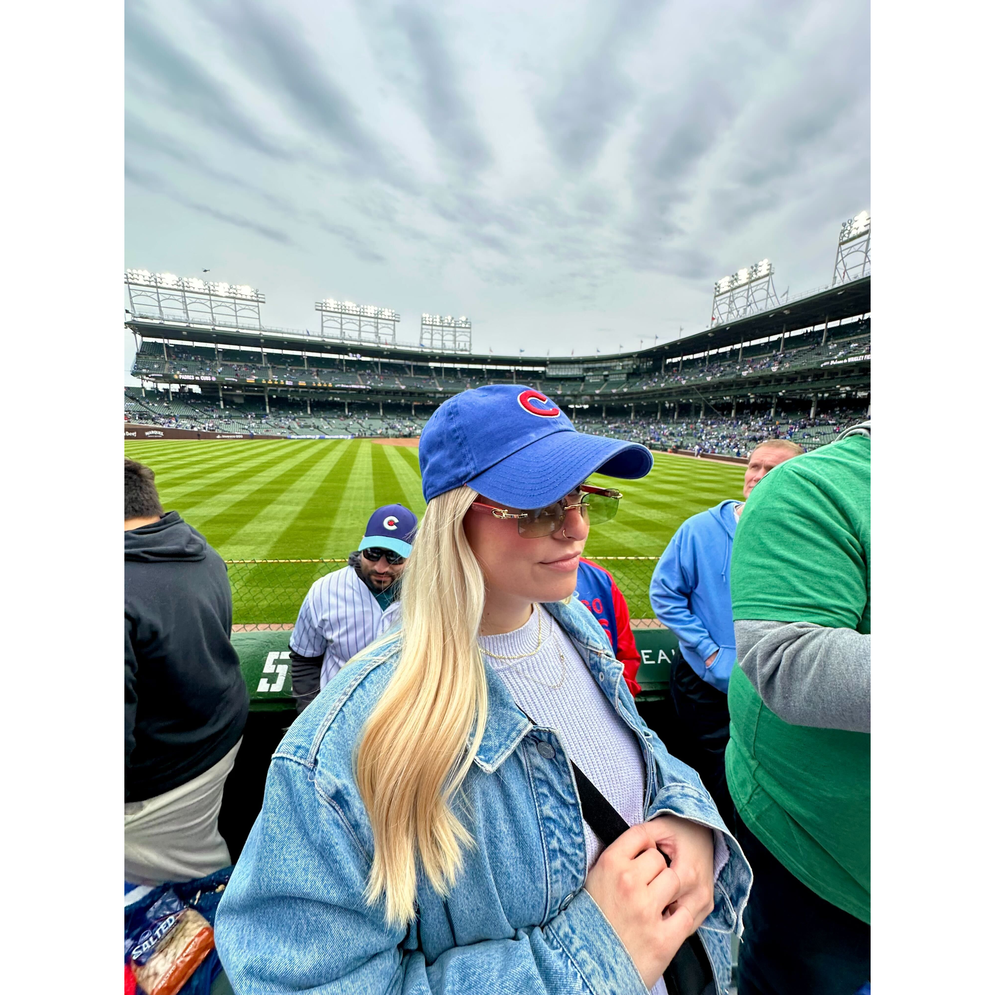 A blonde woman wearing a blue baseball cap, sunglasses and a denim jacket, standing at a baseball stadium