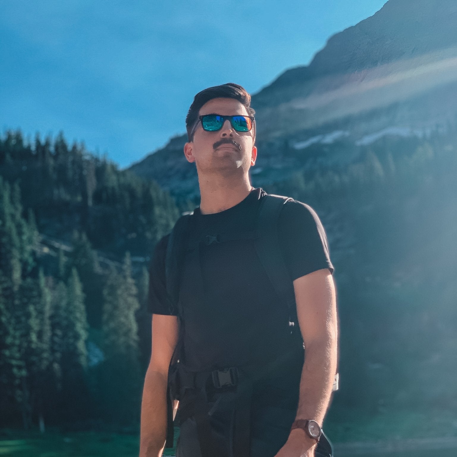 Man wearing reflective sunglasses, black shirt, and backpack standing outdoors with mountain and forest background