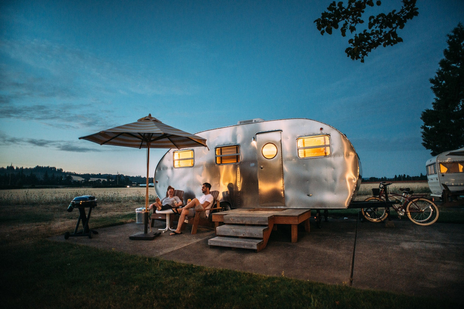 Couple relaxing outside shiny silver camper trailer at dusk with umbrella and barbecue grill