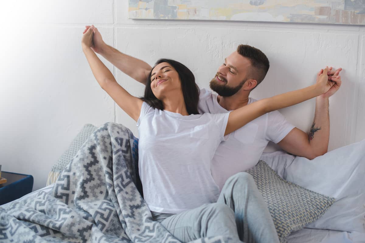 Happy couple in white shirts stretching and relaxing together in bed with cozy patterned blanket in bright bedroom