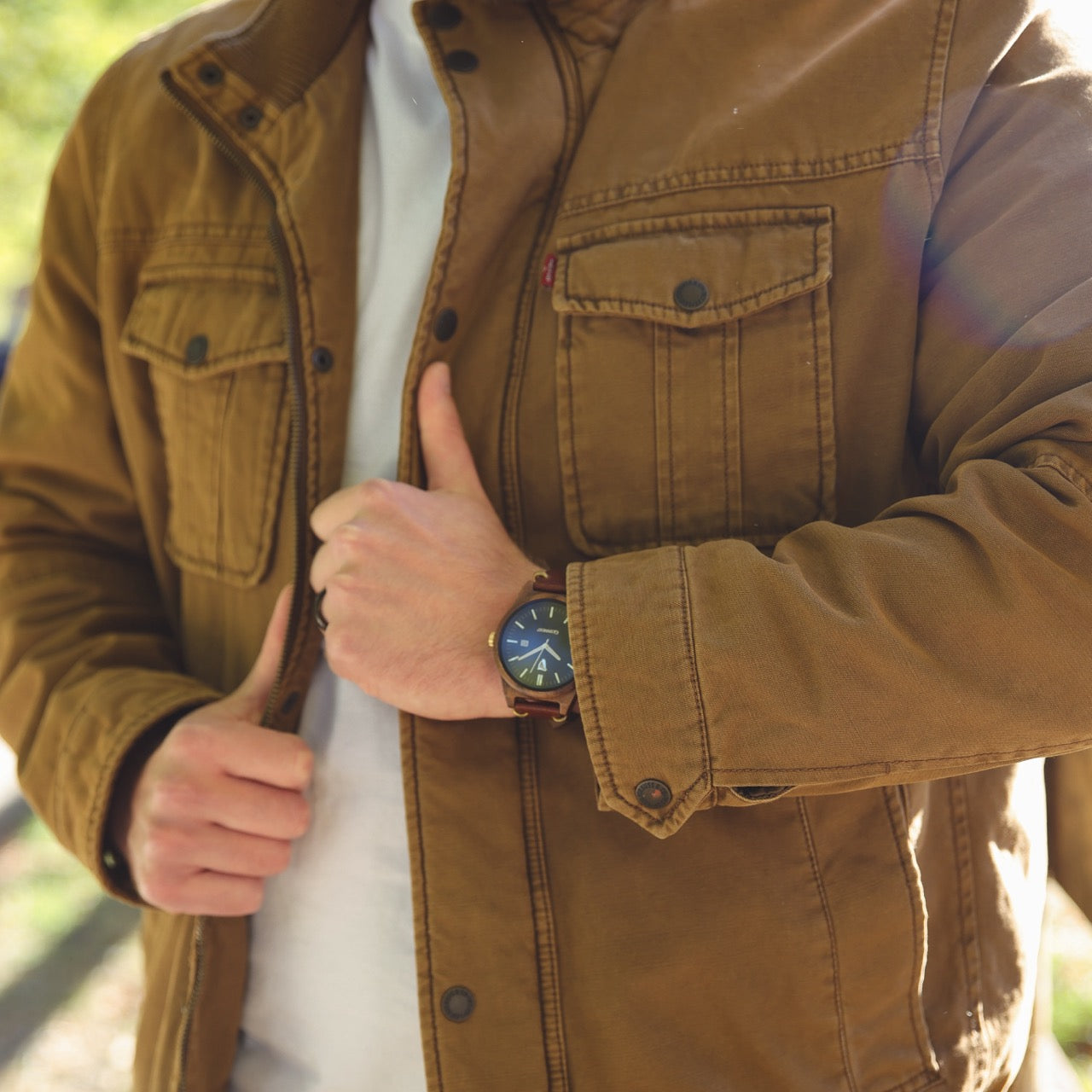 Walnut leather strap watch on wrist with dark dial and gold hands, worn under a jacket.