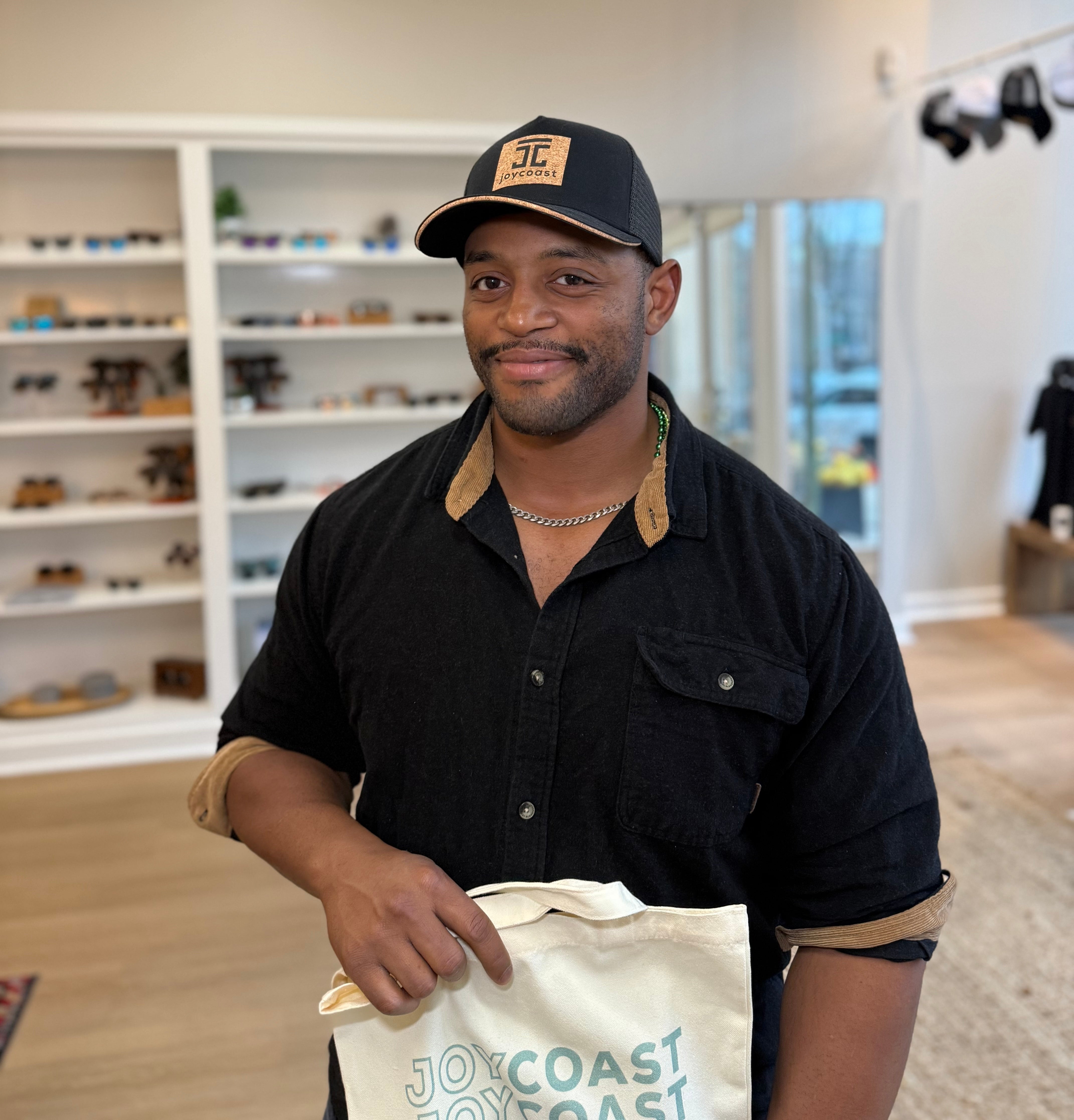 A man wearing a Classic Cork Hat from Joycoast. The hat is a black trucker-style cap featuring a square, sustainable cork patch on the front with the "JC" logo. He is standing in a retail store holding a Joycoast branded canvas tote bag.