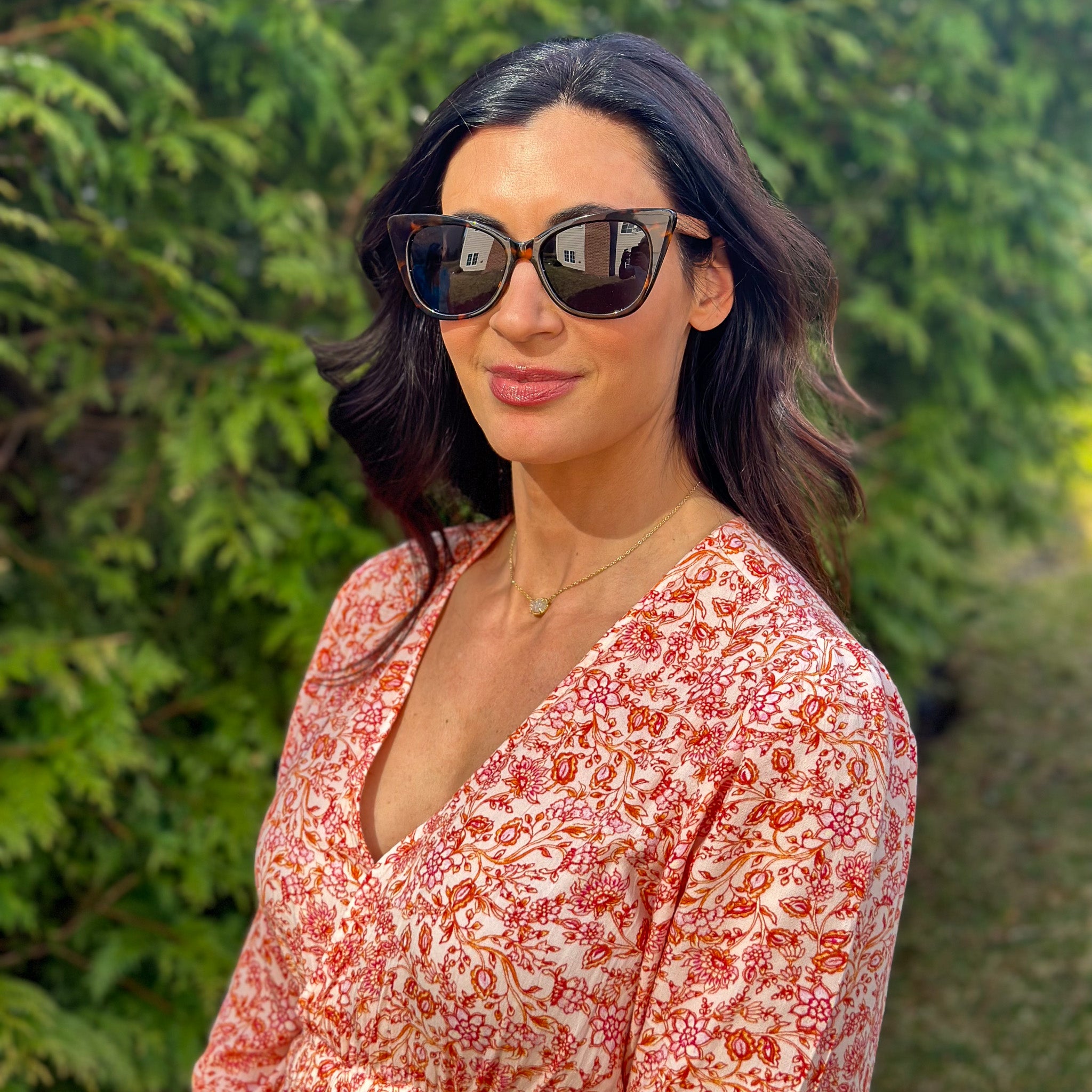 A woman in Joycoast wooden sunglasses and a floral dress stands outdoors, smiling against a bright, sunny background.