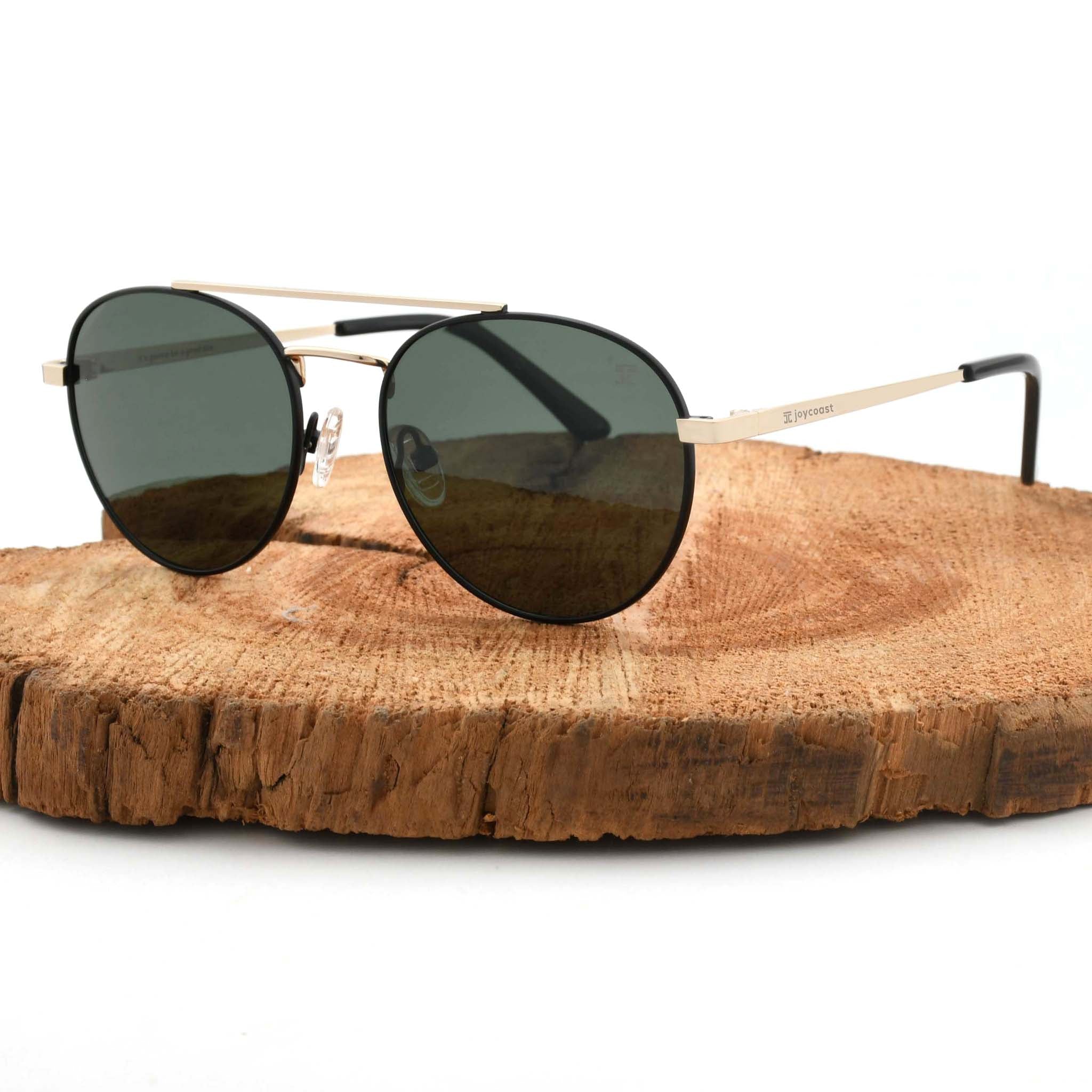 Joycoast Willow round gold sunglasses with dark green lenses, displayed on a natural wood slice.