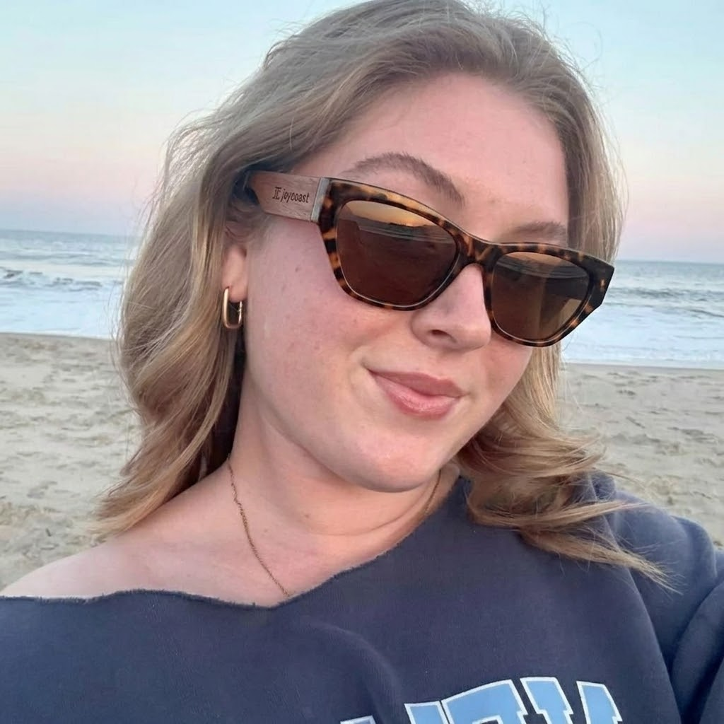 A woman wearing sunglasses smiles while relaxing on the beach, with the ocean and sand in the background.
