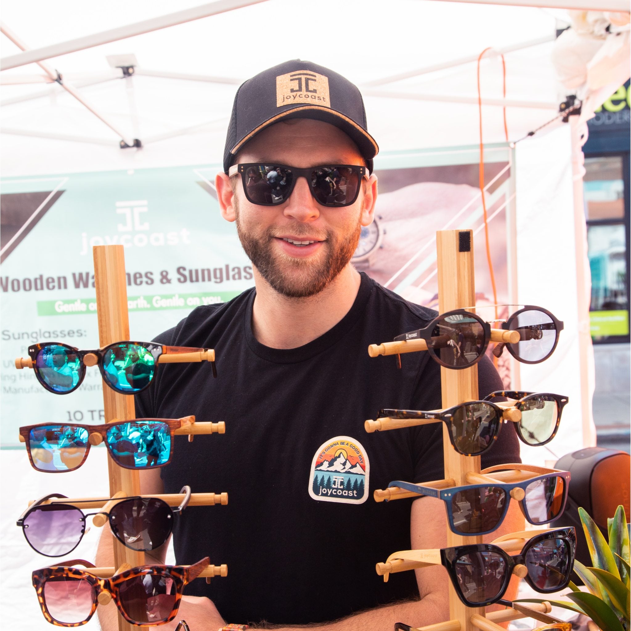 Reese, founder of Joycoast, showcasing a collection of wooden sunglasses at an outdoor event. He is wearing the Classic Cork Hat and branded apparel, standing behind two wooden display stands featuring various eco-friendly eyewear styles, including blue mirrored lenses and tortoiseshell frames.
